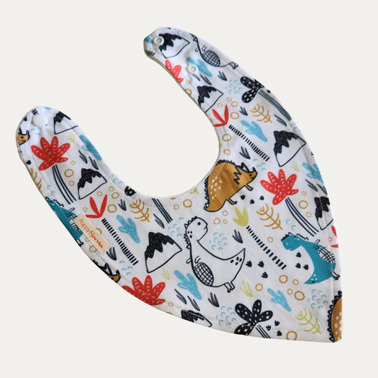 Baby dribble bib with playful teal, mustard and outlined dinosaurs, volcanoes and colourful plants on a cream background.