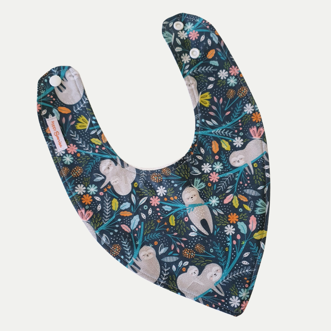 Hangin On baby dribble bib featuring sloths hanging from branches among colourful flowers and foliage on a dark blue background.