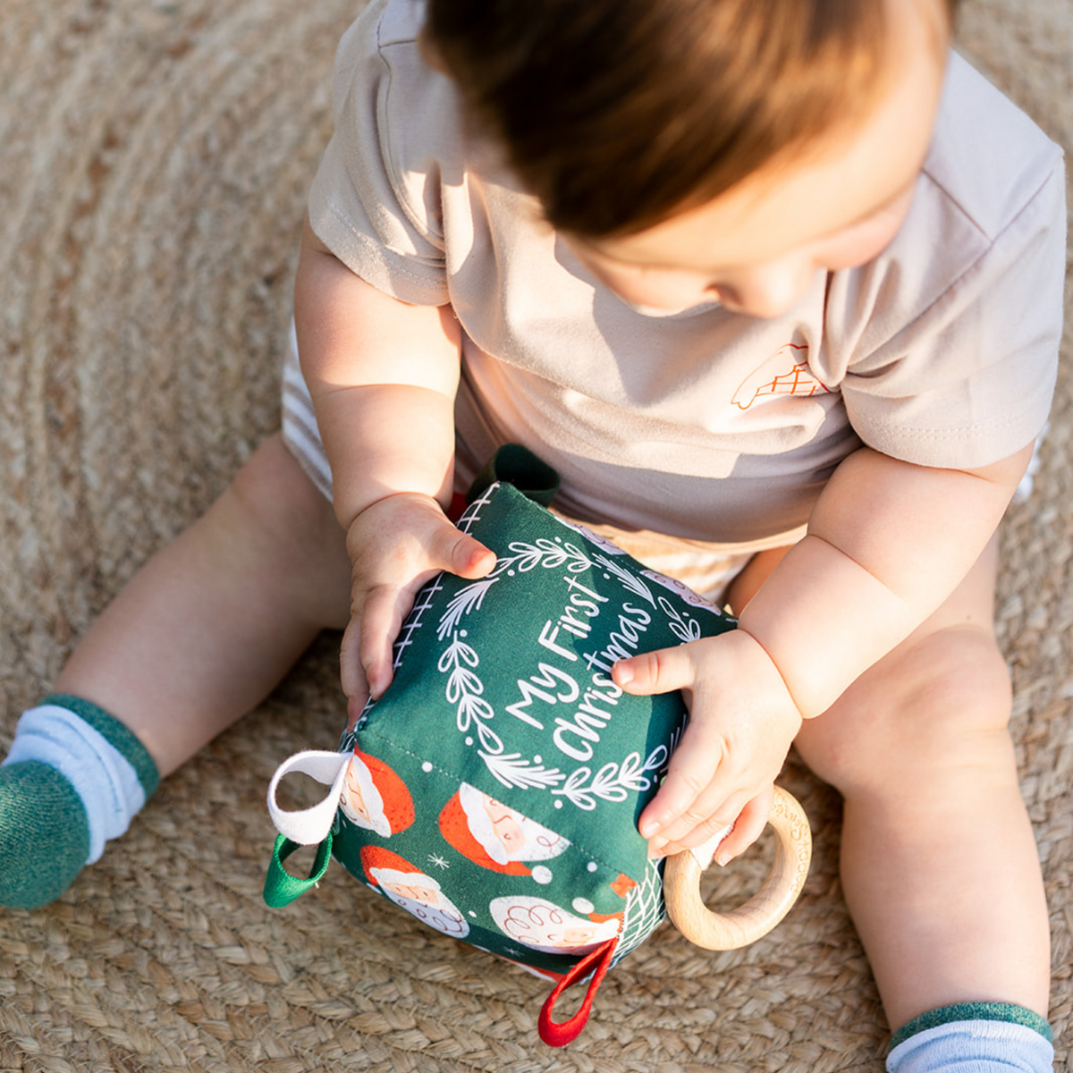 Baby sitting on a jute rug holding the Merry Santa My First Christmas sensory cube with wooden teething ring