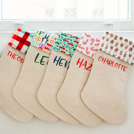 Five personalised linen Christmas stockings with festive print cuffs, hanging in a row along a white window