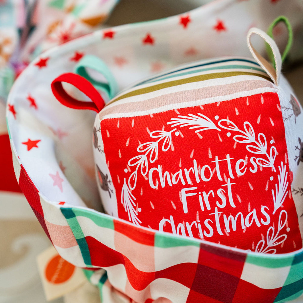 Close-up of a red Charlotte’s First Christmas sensory cube with a white wreath, nestled in a gingham-lined Santa Sack