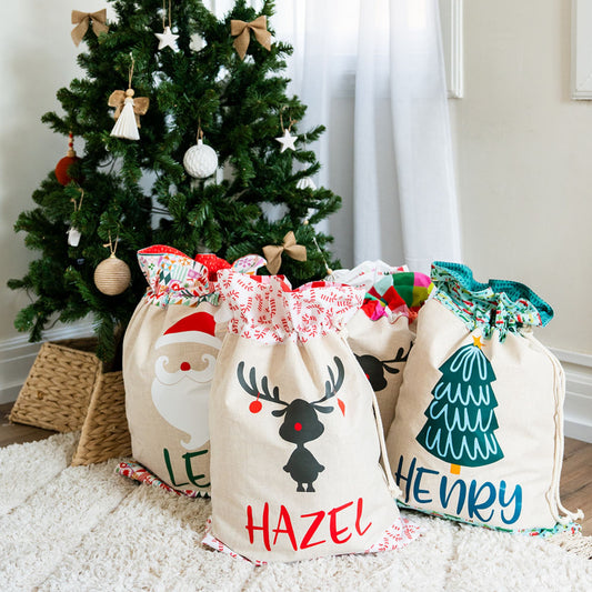 Four personalised linen Santa Sacks with Santa, reindeer and tree designs, gathered under a decorated Christmas tree