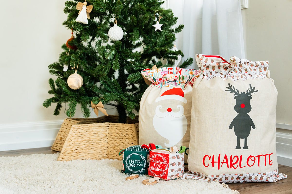 Personalised linen Santa Sacks with Santa and reindeer designs beside a Christmas tree, with My First Christmas sensory cubes in front
