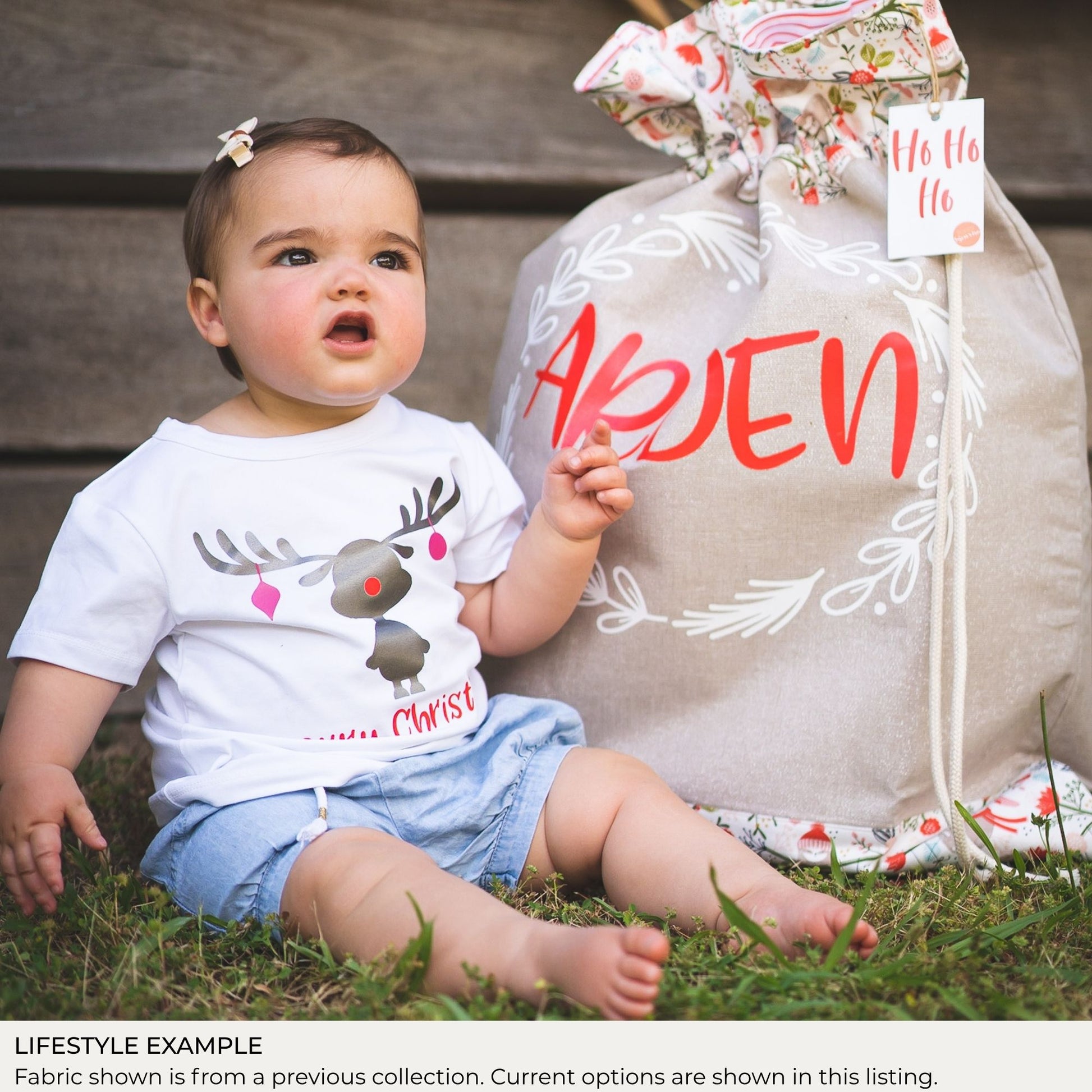 Young child seated beside a personalised Signature Santa Sack outdoors; lifestyle example shows fabric from a previous collection