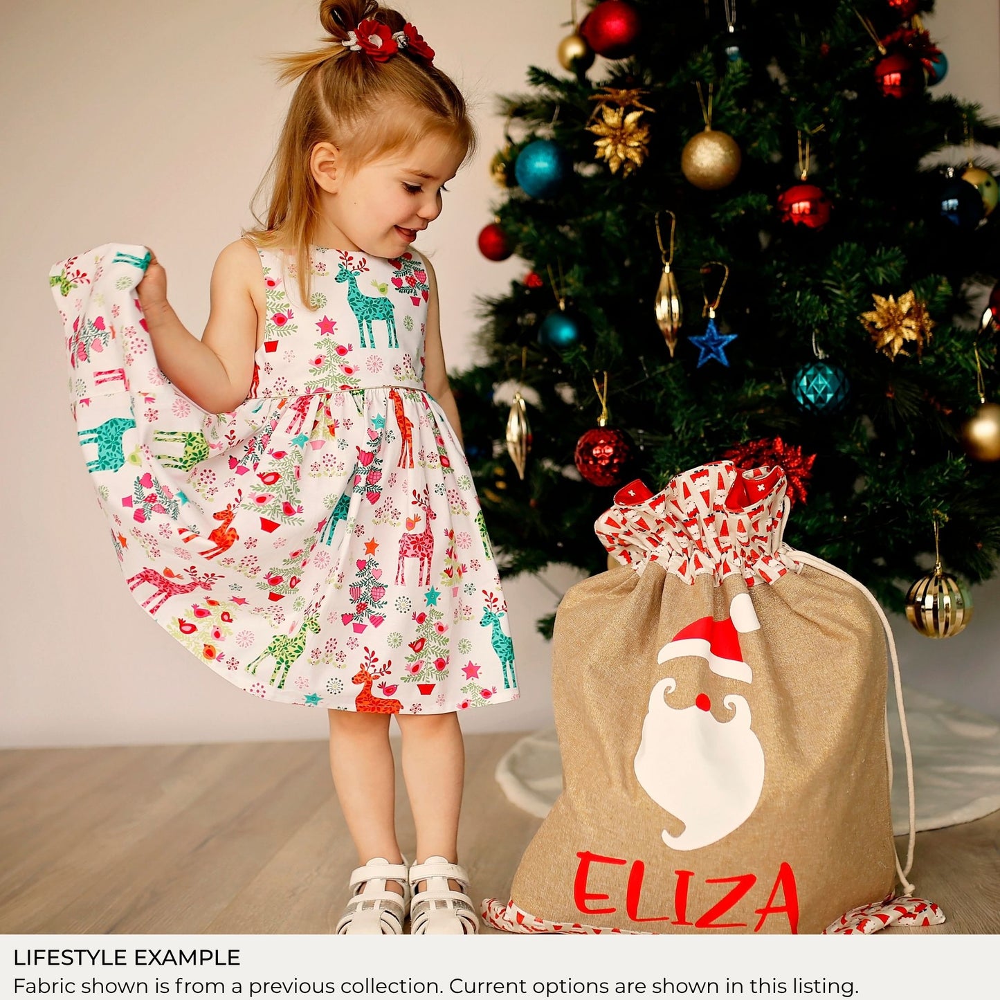 Young girl beside a personalised Signature Santa Sack under a Christmas tree; lifestyle example shows fabric from a previous collection