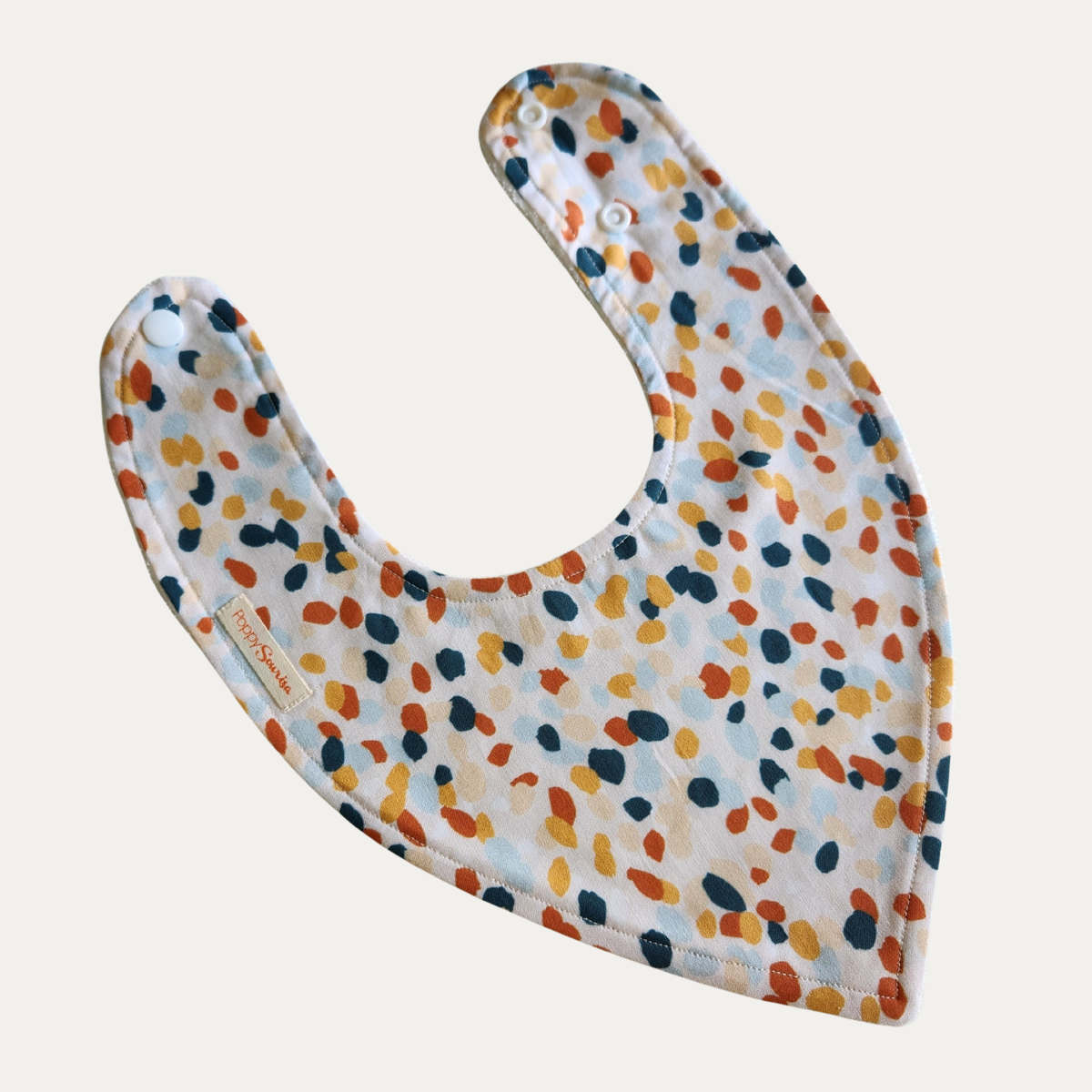 Baby dribble bib with scattered irregular dots in rust, mustard, dark teal, pale blue and beige on a cream background.