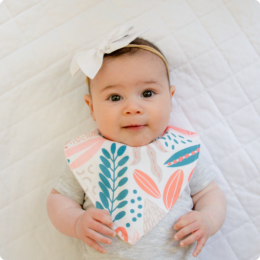Baby wearing a dribble bib with coral, teal and pale grey botanical shapes on a white background.