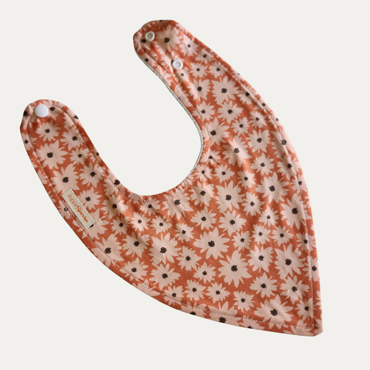 Sunset Daisy baby dribble bib featuring pale pink daisies with dark centres on a warm orange background, with adjustable white snaps.