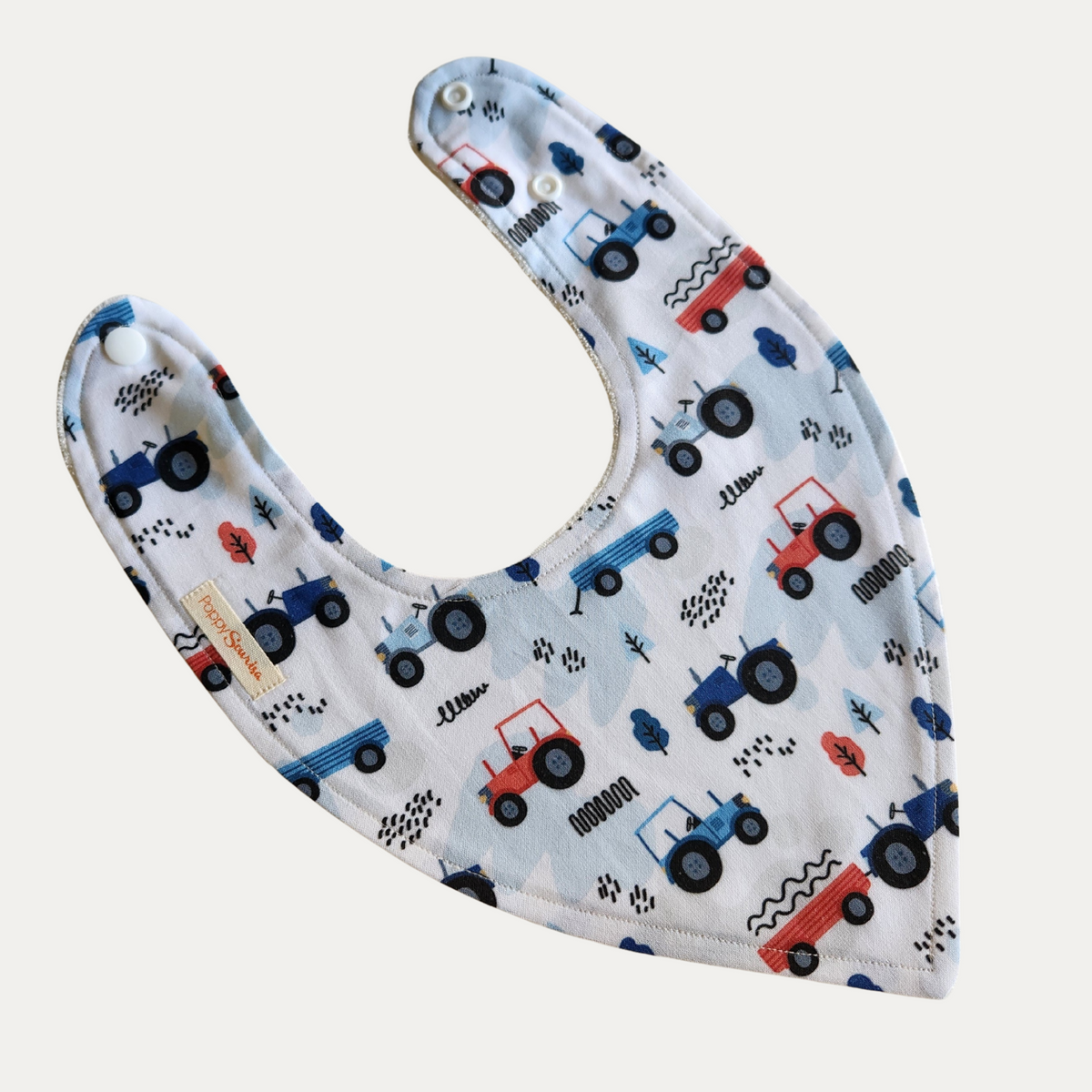 Baby dribble bib with blue and red tractors, trailers and small trees on a pale blue and white background.
