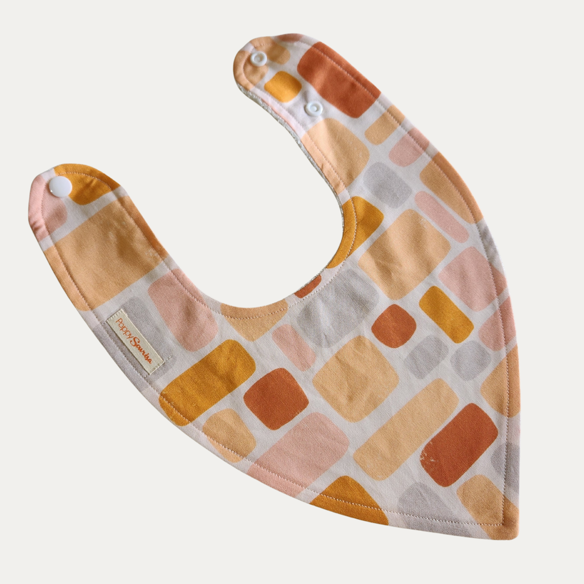 Warm Sand baby dribble bib featuring rounded geometric shapes in rust, mustard, peach, pink and grey on a cream background.